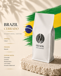 A white bag of CRACK COFFEE BLAERY Brazil Cerrado coffee stands on a textured stone slab against a warm beige background with a green and yellow brushstroke graphic and text describing the coffee varietal, process, and cupping notes.