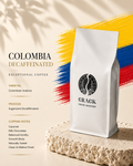 A white bag of Crack Coffee Roastery Colombia Decaffeinated coffee stands on a textured stone slab against a warm beige background with yellow, blue, and red brushstroke accents and product text.