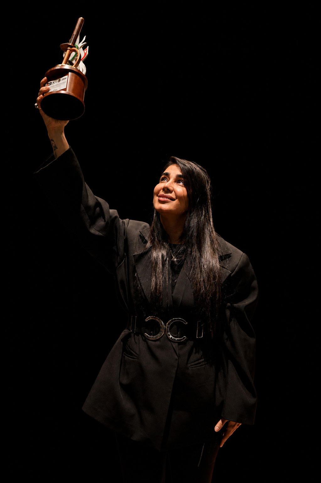 A woman with long dark hair, wearing a black blazer and matching skirt, holds up a bronze trophy against a solid black background.
